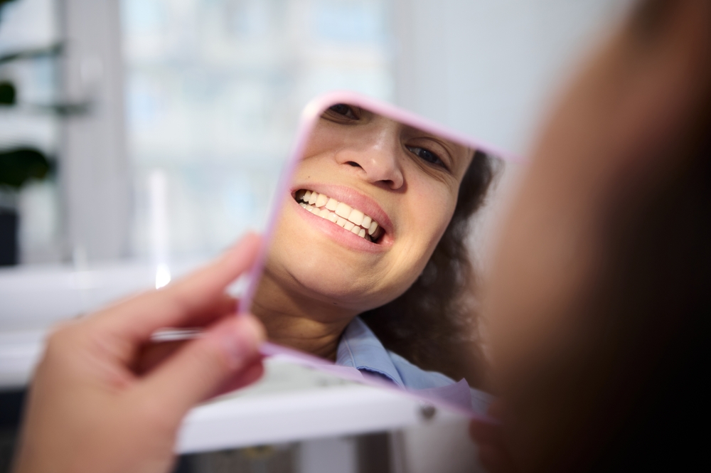 A patient smiling while viewing her teeth in a hand mirror, representing confidence and healthy results from preventive dentistry – Preventive