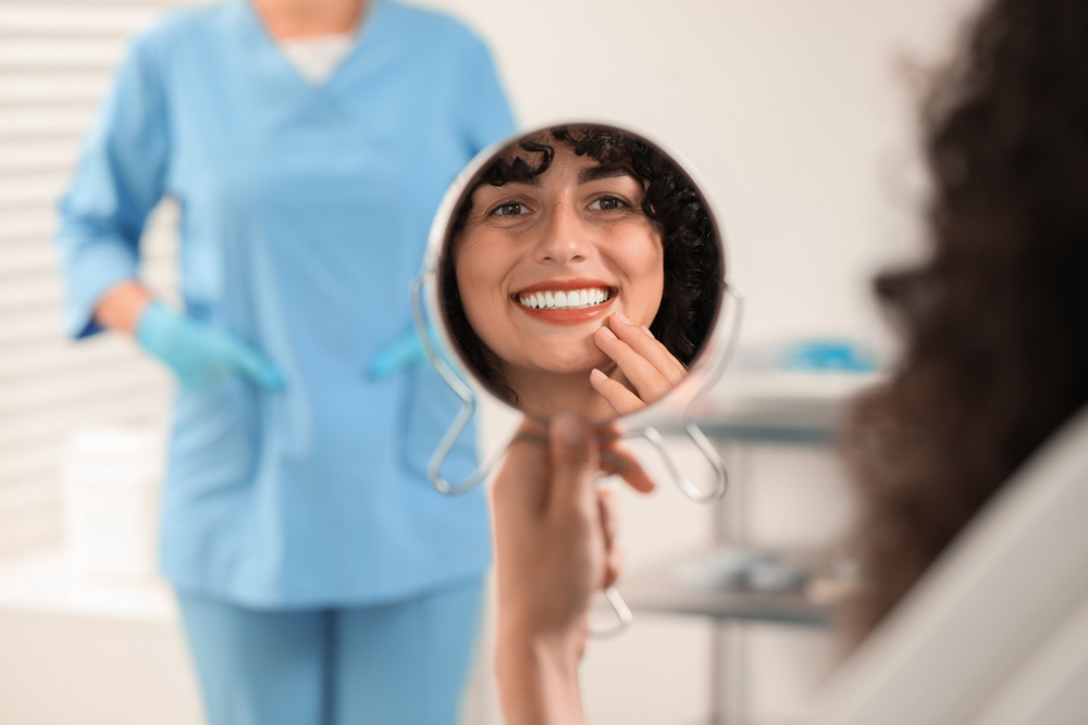 Smile Reveal After Cosmetic Dentistry Treatment – Cosmetic Dentistry A patient smiling at her reflection in a hand mirror, showcasing beautiful results achieved through cosmetic dentistry – Cosmetic Dentistry