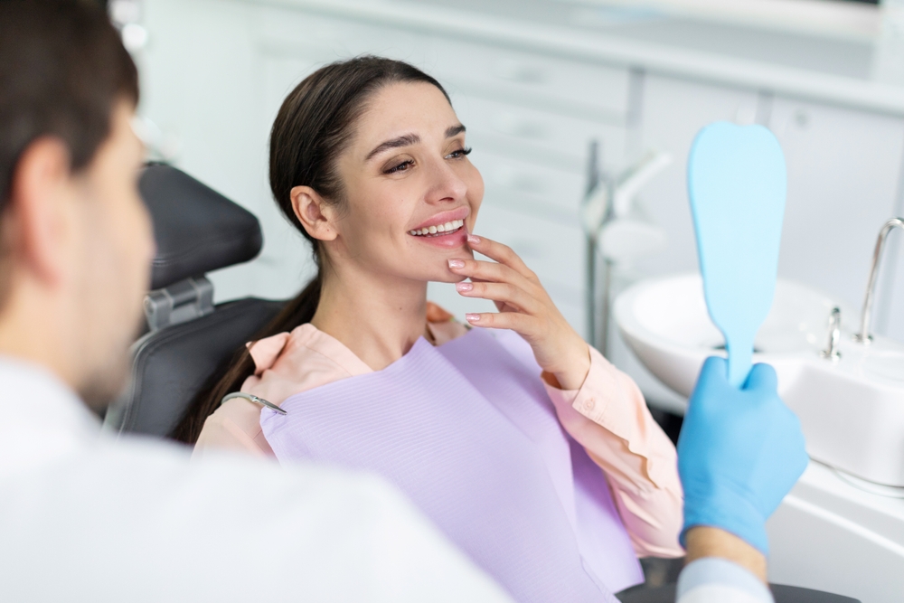 Satisfied Patient Reviewing Smile After Restoration – Restorative Dentistry A patient smiling while examining her teeth in a hand mirror, highlighting confidence and results achieved through restorative dentistry – Restorative Dentistry