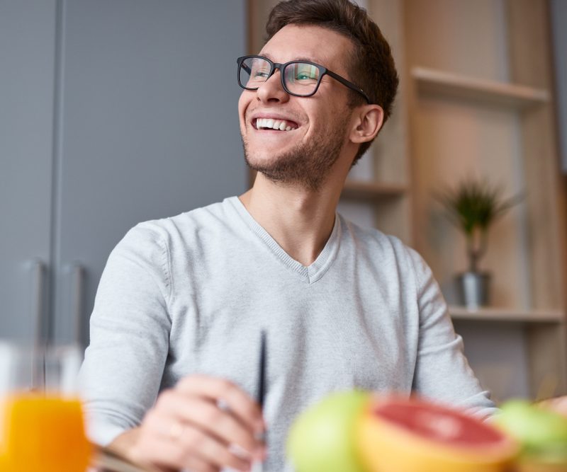 From below of excited handsome man in eyeglasses sitting at desk with fruit and juice smiling away in daylight