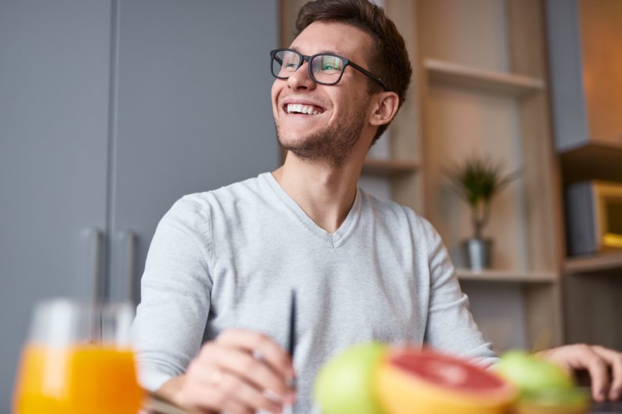 From below of excited handsome man in eyeglasses sitting at desk with fruit and juice smiling away in daylight