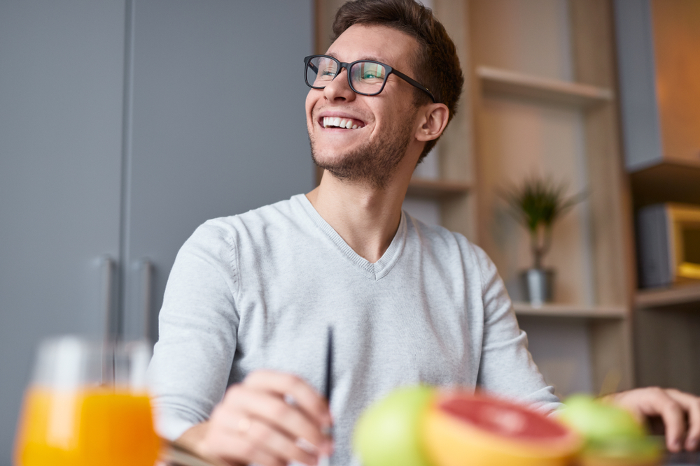 From below of excited handsome man in eyeglasses sitting at desk with fruit and juice smiling away in daylight
