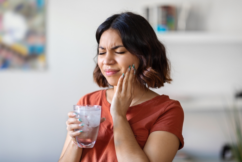 A woman holding her cheek in discomfort while drinking cold water, representing tooth sensitivity or pain that may require emergency dental care – Emergency